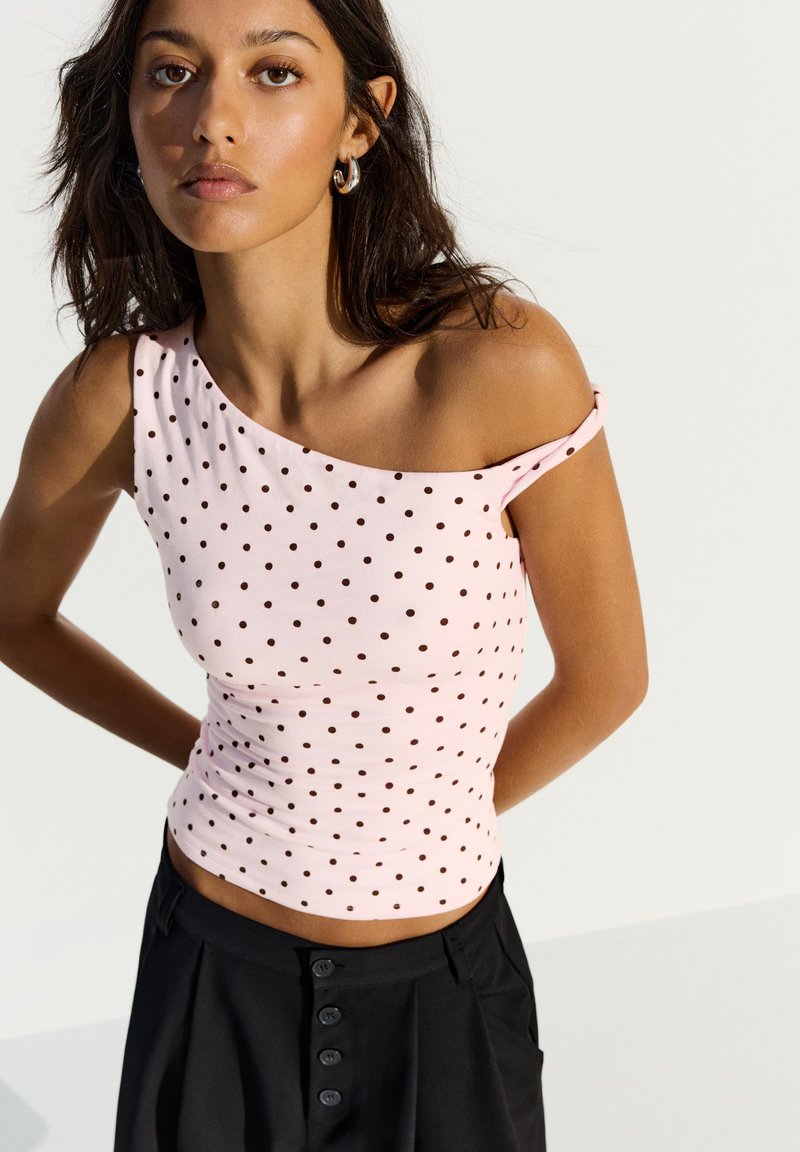 Femme portant un top rose à une épaule avec des pois noirs et un pantalon noir taille haute, debout les mains derrière le dos.