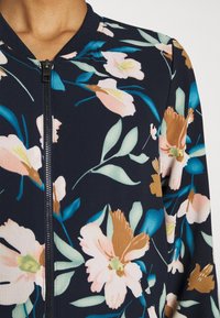 Kvinna som bär en marinblå bomberjacka med dragkedja och ett stort blommönster i ljusrosa, bruna och teal blad och blommor.