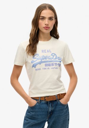T-shirt crème à manches courtes avec un graphique bleu "SuperDry", col rond, en tissu en coton doux, associé à un jean en denim bleu et une ceinture marron.
