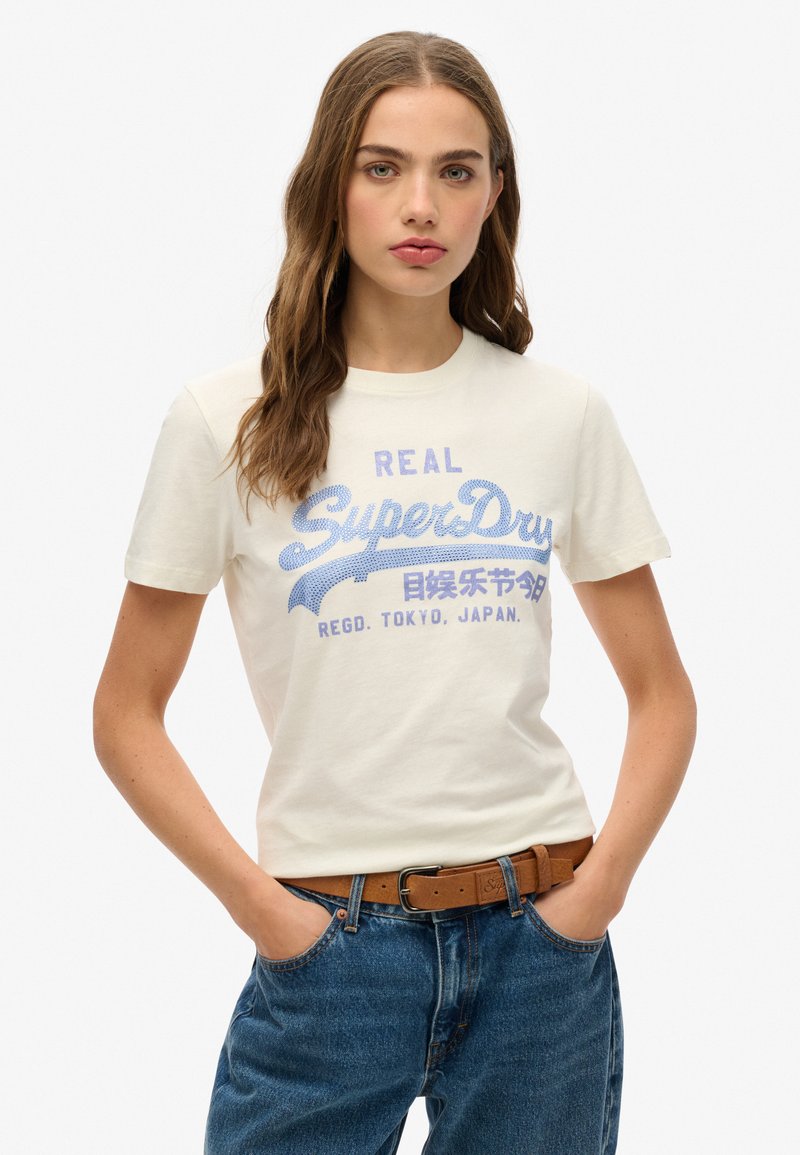 T-shirt crème à manches courtes avec un graphique bleu "SuperDry", col rond, en tissu en coton doux, associé à un jean en denim bleu et une ceinture marron.