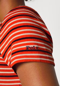 Červenobílý pruhovaný bavlněný košile s námořnickými modrými akcenty. Obsahuje logo s nápisem "Polo" na rukávu. Krátké rukávy a žebrovaná textura.