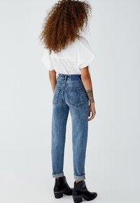 Jeans de mezclilla azul de cintura alta, ajuste entallado, con puños doblados y bolsillos parche, complementados por unas botas negras de cuero hasta el tobillo.