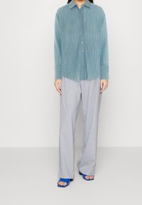 Jasnoniebieska, prążkowana koszula z długim rękawem i zapięciem na guziki, zestawiona z luźnymi, szarymi spodniami oraz niebieskimi sandałami na obcasie. Widoczne detale teksturowego materiału.