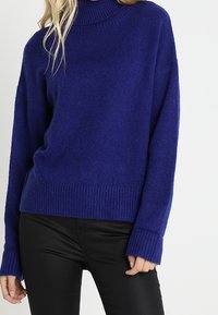 Maglione blu lavorato a maglia con collo alto, polsini e orlo a coste. Texture morbida, vestibilità comoda e maniche di lunghezza media, abbinato a pantaloni neri.