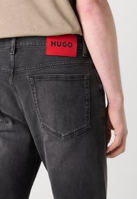 Black denim jeans feature a prominent red tag with "HUGO" on the waistband, with five-pocket design and a textured finish.