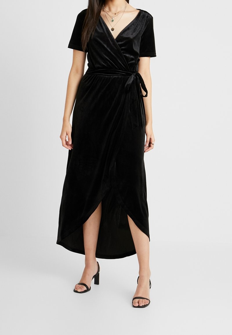 Schwarzes Samt-Wickelkleid mit kurzen Ärmeln, V-Ausschnitt, Bindebändern in der Taille und einem hohen Schlitz vorne. Mit Absatzsandalen wird der Look abgerundet.