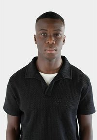 Svart kortärmad pikétröja med strukturerad mönstring och inskuren krage, bärs över en vit t-shirt med rund hals.