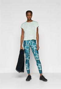 Ανοιχτό μέντας πράσινο κοντό μπλουζάκι με γεωμετρικό σχέδιο, συνδυασμένο με τιρκουάζ κολάν με λευκές αφηρημένες μορφές και μαύρα αθλητικά παπούτσια.