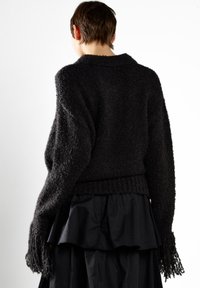 Osoba z krótkimi włosami, ubrana w czarny sweter o teksturowanym wzorze z frędzlami na rękawach oraz warstwową czarną spódnicą, zwrócona plecami w kierunku białego tła.