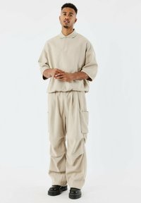 Polo shirt beige di grandi dimensioni con colletto e maniche corte, abbinato a pantaloni a vestibilità rilassata con due tasche laterali e un tessuto testurizzato.