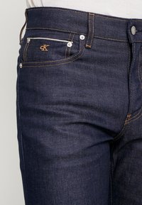 Tummansiniset farkut, joissa sileä rakenne, kullanväriset saumat, pieni CK-logo ja hopeanväriset metalliosat etutaskuissa.