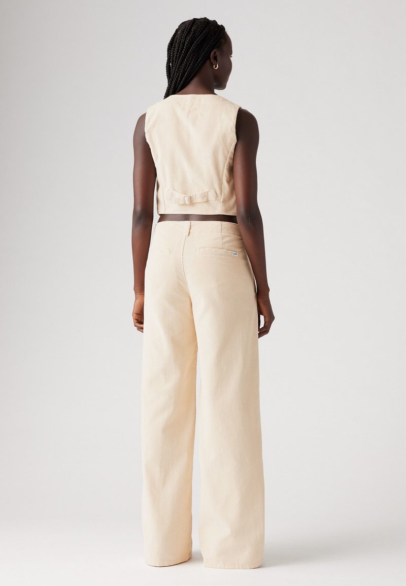 Beige Cropped-Weste mit einer Schleifenverzierung auf der Rückseite, kombiniert mit hochtaillierten, locker sitzenden beigen Hosen mit zwei Gesäßtaschen.
