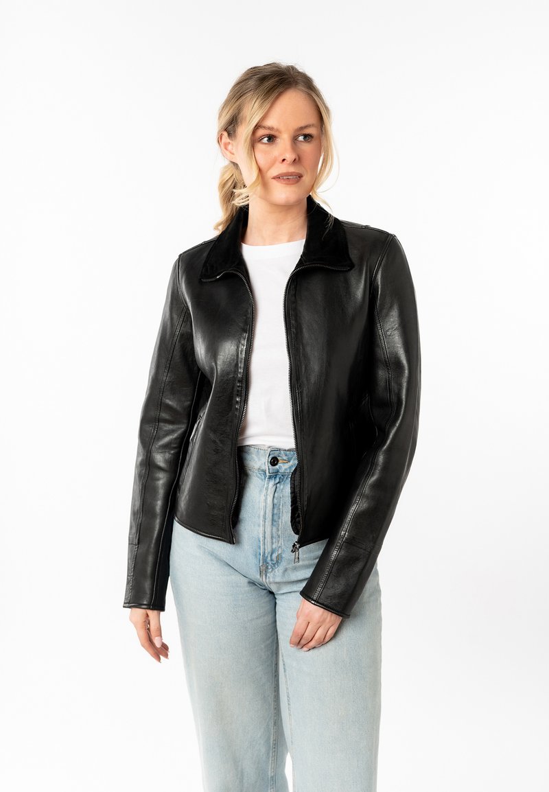 Zwarte leren jas met een rits aan de voorkant, puntige kraag en gestroomlijnd ontwerp, gecombineerd met lichtblauwe jeans en een wit topje.