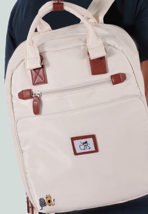 Cremefarbener Rucksack mit braunen Lederelementen, kleinem Reißverschlussfach vorne und Cartoon-Katzen-Patches in der unteren Ecke sowie auf dem mittleren Etikett.