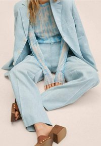 Costume en lin bleu clair avec pantalon à jambes larges et veste cintrée. En dessous, un haut ajusté présente un motif tie-dye bleu. Sandales marron à talons blocs.