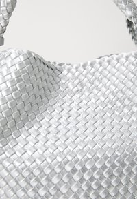 Ασημένια τσάντα χειρός από πλέγμα με υφή που διαθέτει ξεχωριστό σχέδιο και στρογγυλό σχήμα. Περιλαμβάνει μια μικρή κυκλική λεπτομέρεια.