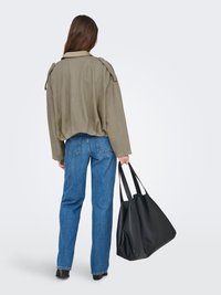 Khakifarget jakke med skulderklaffer, kombinert med blå jeans med rette ben og en stor svart handlenett som bæres av brukeren.