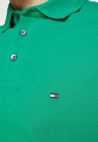 Polo-shirt vert en tissu texturé, avec un col, trois boutons et un petit logo brodé en rouge, blanc et bleu.