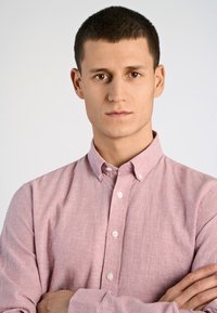 Lichtroze button-up shirt met een kraag, een gladde textuur en een subtiel geweven patroon. Model heeft kort haar en heeft de armen gekruist.