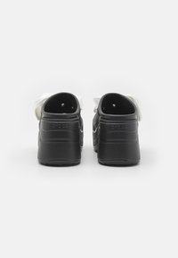 Black clog-style shoes with a textured upper, smooth finish, and soft white accents. Features a low heel and open back design.