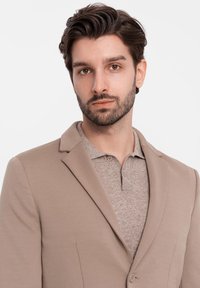 Beige Blazer aus weichem Stoff, mit Reverskragen und einem Knopfverschluss, getragen über einem hellbraunen Poloshirt mit zwei Knöpfen.