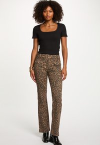 Vrouw met krullend haar, gekleed in een zwart T-shirt met korte mouwen, panterprint broek met voorzakken en zwarte enkellaarsjes, staand tegen een witte achtergrond.