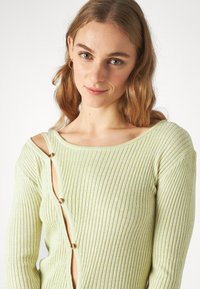 Lys grønn ribbestrikket genser med rund hals og lange ermer, med en sideluke som festes med brune knapper.
