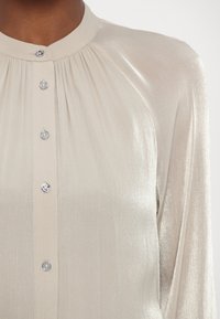 Blouse beige clair à manches longues, avec un devant plissé et un col rond. Dotée d'un tissu brillant et de boutons argentés le long du centre.