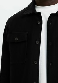 Mustragen, musta, mis on kaunistatud rinna taskuga, kantud valge ümmarguse kaelusega T-särgi peal, nähtav kaelast kuni keskpuusani.