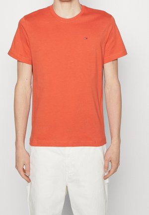 T-shirt orange en coton à manches courtes avec un col rond, comportant un petit logo sur la poitrine, associé à un pantalon blanc.