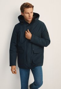 Granatowa parka z kapturem, wyposażona w przednie kieszenie oraz kieszeń na rękawie na suwak, wykonana z teksturowanego, wodoodpornego materiału.