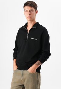 Ung mand i en sort lynlås-trøje står med hænderne i lommerne, iført lyse jeans og en rosatattoo på sin underarm.