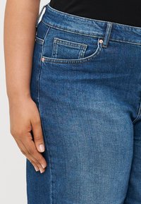 Sötét kék farmernadrág sima textúrával, öt zsebbe, varrott varratokkal és közepes derekú derékkal.