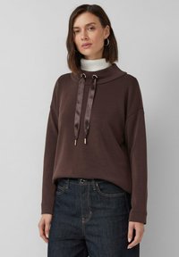 Femme aux cheveux bruns courts portant un sweat à capuche marron foncé avec un large col et des cordons en satin, un jean foncé et des boucles d'oreilles créoles sur fond uni.