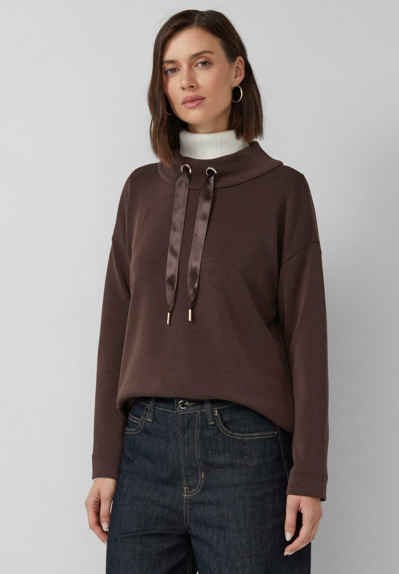 Femme aux cheveux bruns courts portant un sweat à capuche marron foncé avec un large col et des cordons en satin, un jean foncé et des boucles d'oreilles créoles sur fond uni.