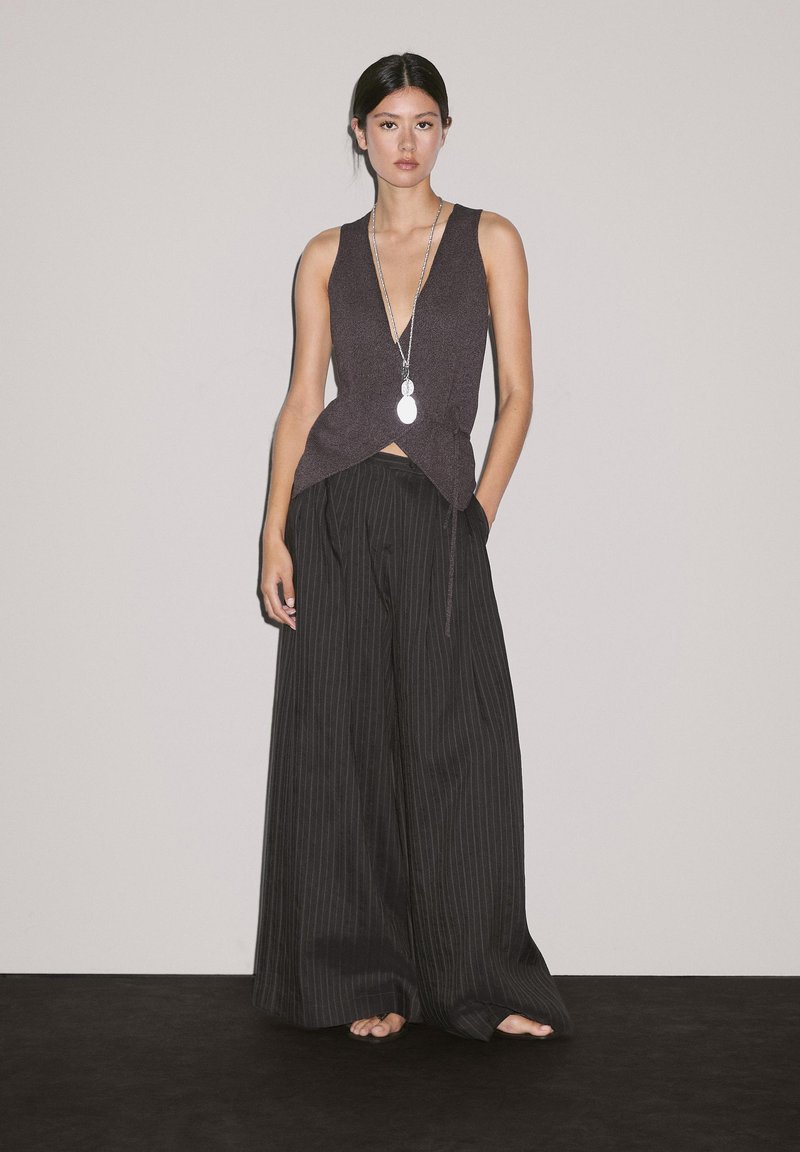 Top sans manches marron à décolleté en V profond associé à un pantalon large à rayures noires. Le modèle porte un collier avec pendentif circulaire.