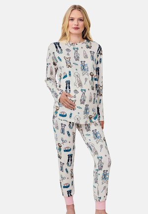 Kvinde iført hvid langærmet top og bukser med farverige illustrationer af hunde og kæledyrsudstyr, stående med den ene hånd på maven.