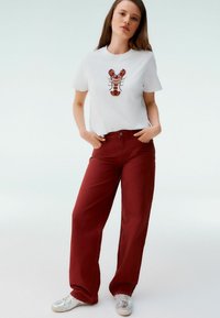 Maglietta bianca in cotone con ricamo di aragosta rossa, abbinata a pantaloni bordeaux a vita alta e sneaker argentate. Design semplice, look casual.