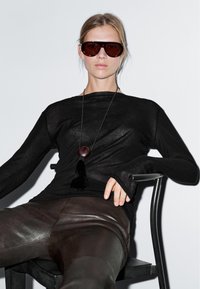 Kvinde iført oversize brune solbriller, sort langærmet top, læderbukser og en lang halskæde med en stor vedhæng, siddende på en stol.