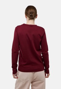 Chemise bordeaux à manches longues avec col rond, réalisée en tissu doux. Présente une texture lisse et un design simple, portée avec un pantalon de couleur claire.