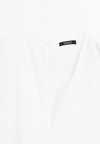 Witte blouse met een V-hals, gemaakt van lichte stof. Bevat een discrete achterrits en een zwart merketiket bij de nek.