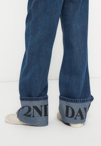 Τζιν παντελόνια με φαρδιά μπατζάκια σε σκούρο μπλε πλύσιμο, με μεγάλα αναδιπλωμένα ρεβέρ που εμφανίζουν τη φράση "2ND DAY" με έντονα μαύρα γράμματα.