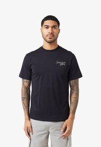 Rövid hajú, tetovált férfi fekete pólót visel, amelyen fehér felirat található a mellkasán, valamint világosszürke rakott nadrágot, és fehér háttér előtt áll.