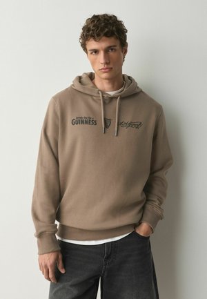 Young man with curly hair wearing a light brown hoodie with "Lovely day for a Guinness" and graphic text, paired with dark jeans.