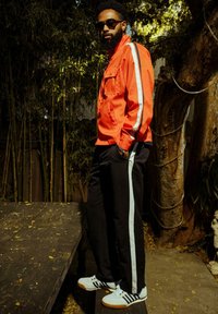 Veste zippée rouge avec des rayures blanches sur les côtés, pantalon de jogging noir avec des rayures bleu clair, et chaussures de sport blanches avec des accents noirs.