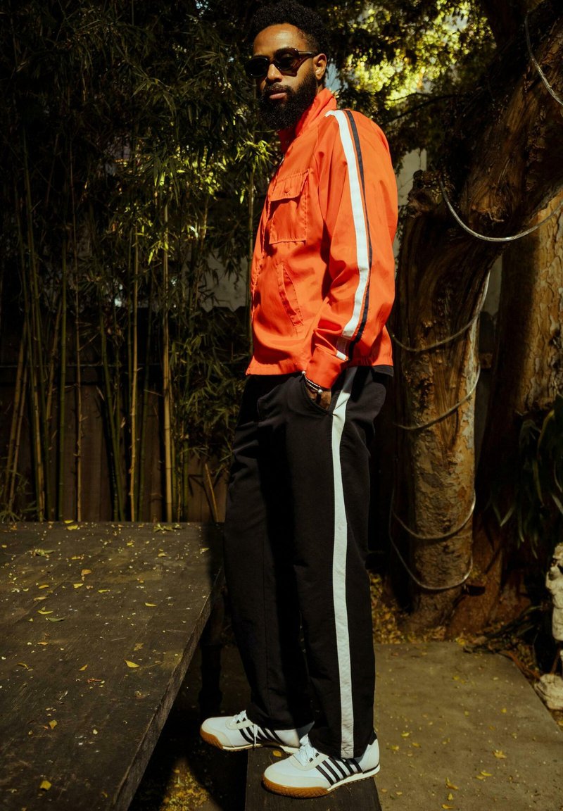 Veste zippée rouge avec des rayures blanches sur les côtés, pantalon de jogging noir avec des rayures bleu clair, et chaussures de sport blanches avec des accents noirs.