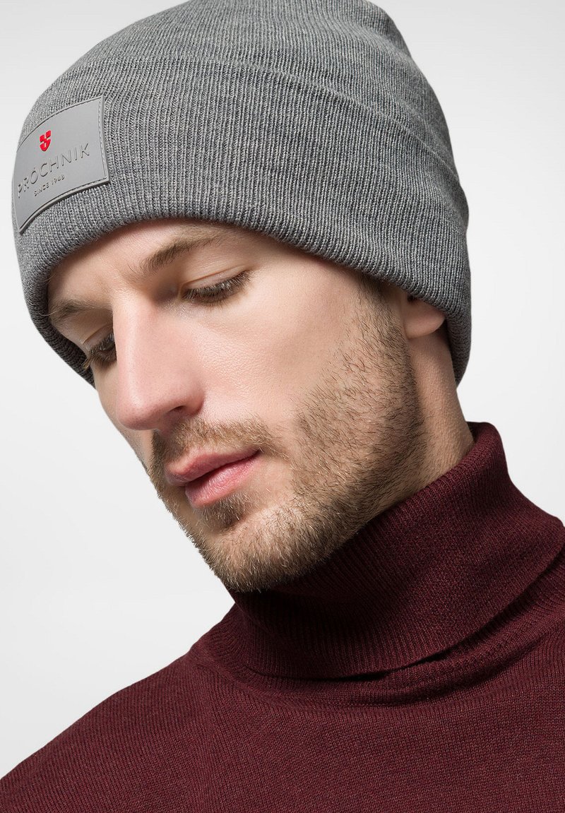 Szary dzianinowy beanie o prążkowanej teksturze, z naszytą metką z logo. Noszone z bordo golfem, podkreślającym design odzieży.