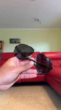 Μαύρα γυαλιά ηλίου με σκούρους φακούς, λεπτό μεταλλικό σκελετό και υφασμάτινα πλαστικά χερούλια, κρατημένα σε ένα χέρι μπροστά από φόντο κόκκινας καναπέ.