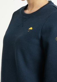 Sweatshirt bleu marine en tissu doux, avec un col rond, des poignets côtelés et une petite broderie de parapluie jaune sur la poitrine.