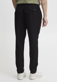 Pantalon en lin noir à coupe décontractée, doté de deux poches arrière et d'une texture lisse. Porté avec des baskets blanches et un haut vert.